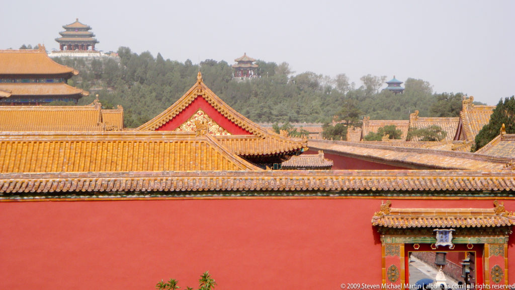 Layered yellow-tiled palace roofs above red walls in the Forbidden City, with wooded Jingshan hill in the distance.