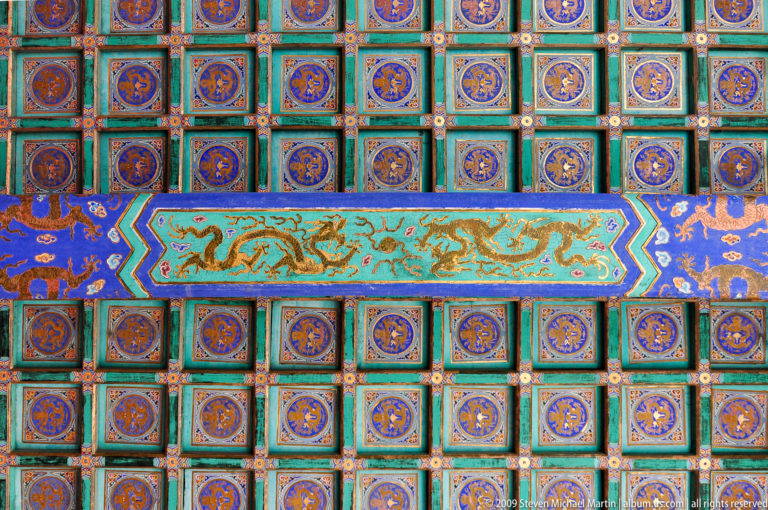 Upward view of a polychrome coffered ceiling in the Forbidden City with a central turquoise dragon panel surrounded by repeating square coffers.