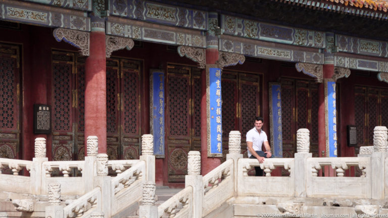 Male visitor seated on a white stone balustrade in front of a palace hall with red columns and blue calligraphy plaques in the Forbidden City.