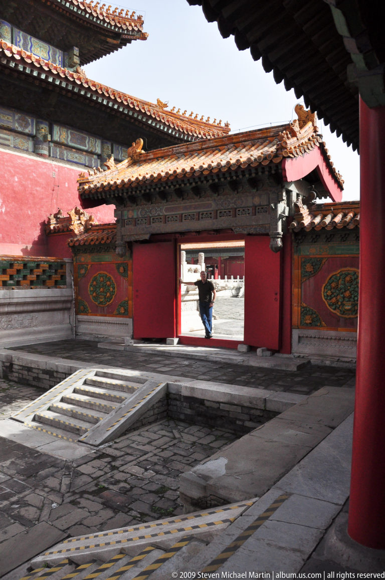 Distant full-body view of a male visitor centered in an open red doorway at the Forbidden City, framed by decorated wall panels and a stone-lined channel in the foreground.