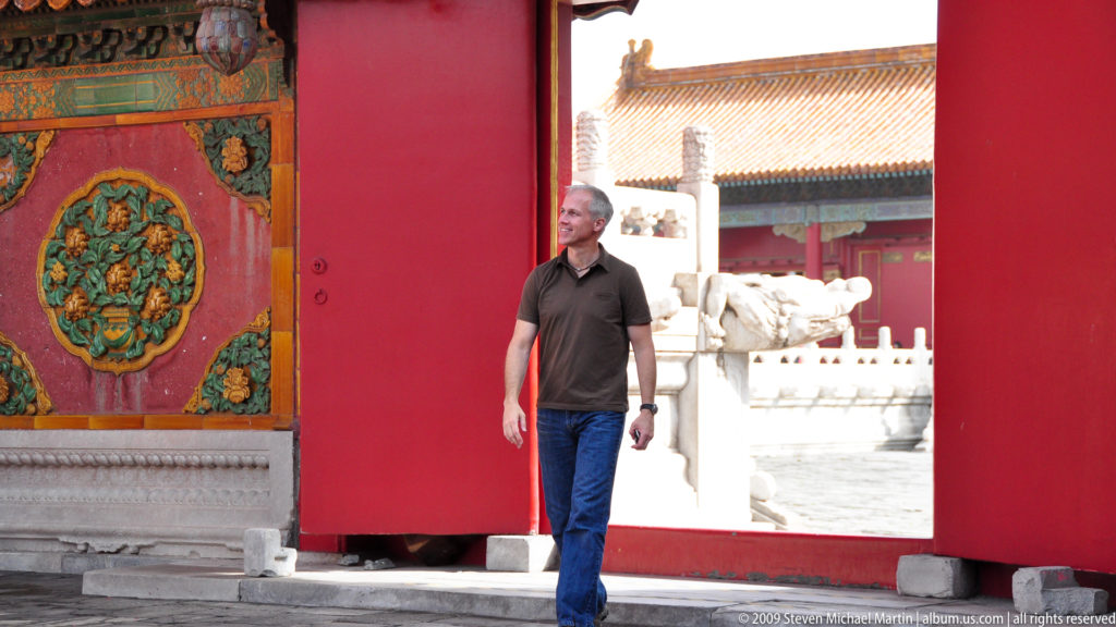 Male visitor mid-stride walking through a red palace gateway in the Forbidden City, with a carved dragon-head waterspout visible in the bright courtyard beyond.