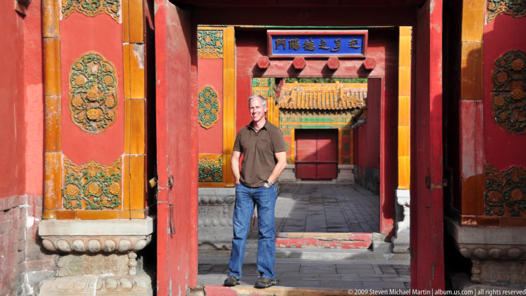 Full-length portrait of a male visitor standing slightly left of center between aligned red-and-gold palace doorways in the Forbidden City, with a blue calligraphy plaque above the inner gate.