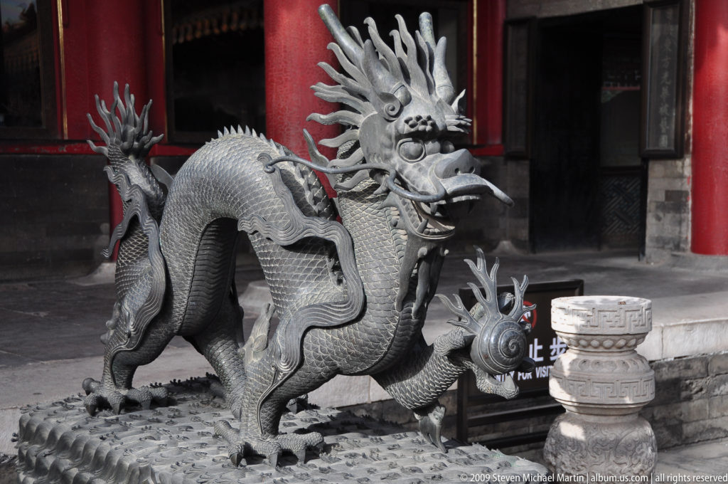 Cast-metal dragon sculpture with a long scaled body and antler-like horns standing on a textured base in a Forbidden City palace courtyard before red columns.