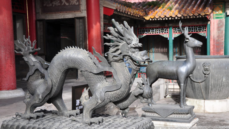 Cast-metal dragon sculpture with antler-like horns and a long scaled body standing on a textured base before red palace columns in the Forbidden City courtyard.