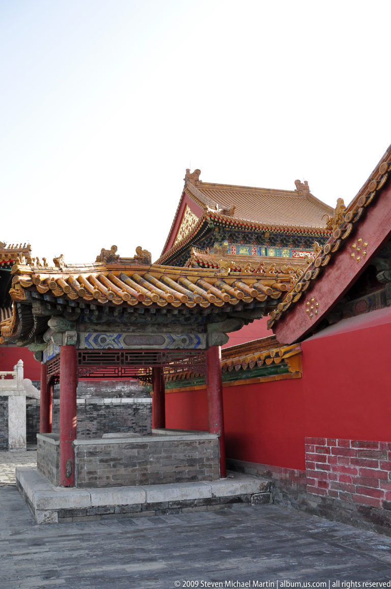 Red-columned corner pavilion with a painted lintel beneath overlapping yellow-tiled roofs in the Forbidden City.