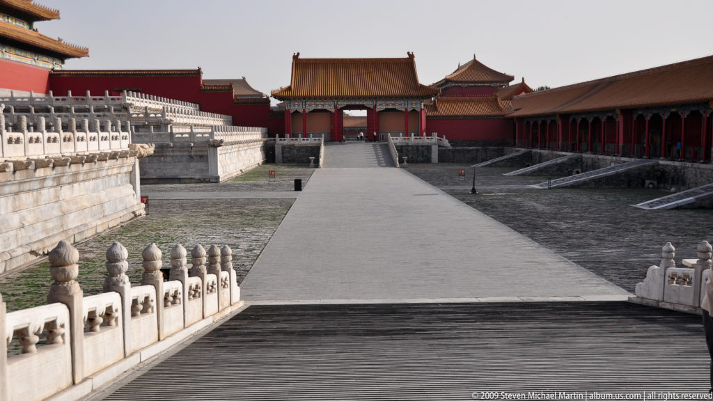 Axial view along a wide stone courtyard toward successive red-and-yellow palace gates in the Forbidden City.