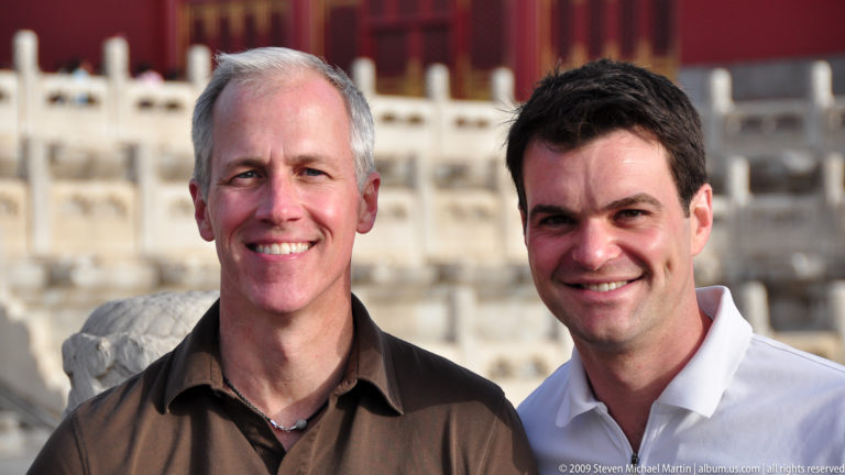 Two smiling male visitors in a close portrait with white carved palace balustrades softly blurred in the background.