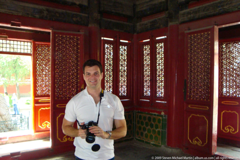 Male visitor holding a camera inside a red-lacquered pavilion with red lattice doors and green lower panels in the Forbidden City.