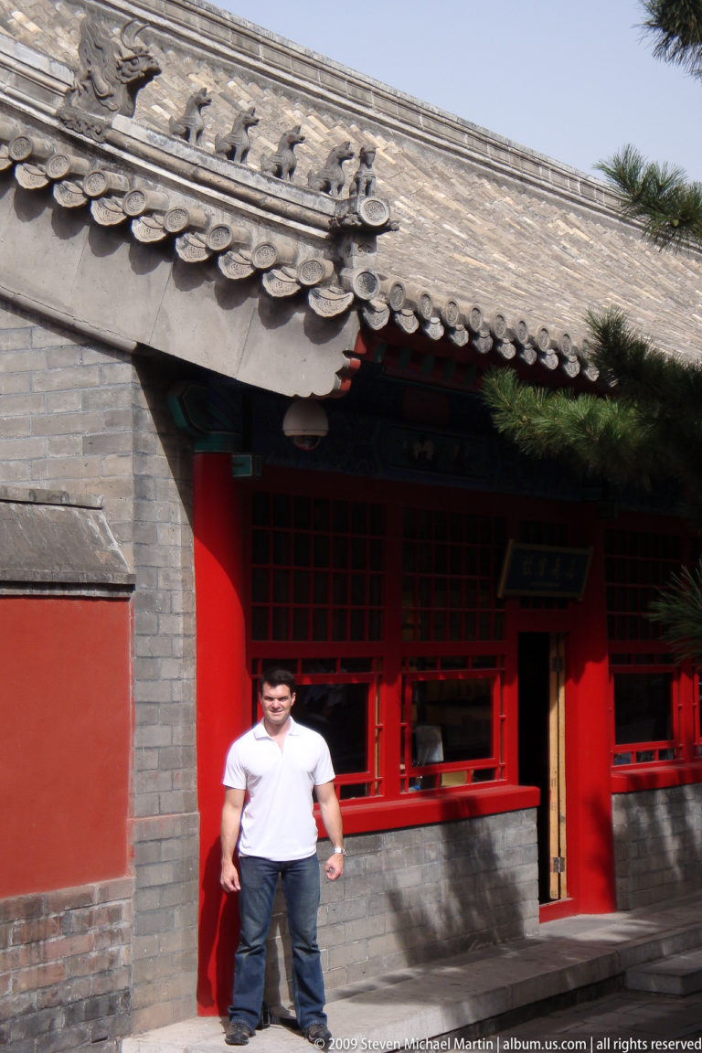 Male visitor standing beneath gray-tiled eaves at the entrance of a red-latticed palace building in a secondary courtyard.