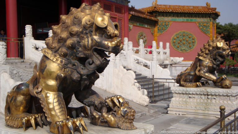Side view of a gilt-bronze male guardian lion pressing a patterned brocade ball, with a second guardian lion visible across the palace entrance.