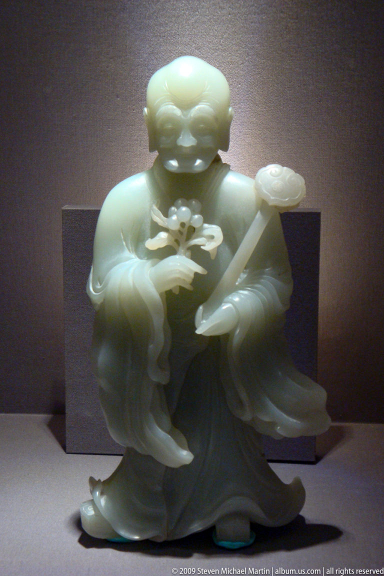 Translucent pale-green jade female figure in long robes, holding a blossom and long implement in a display case.
