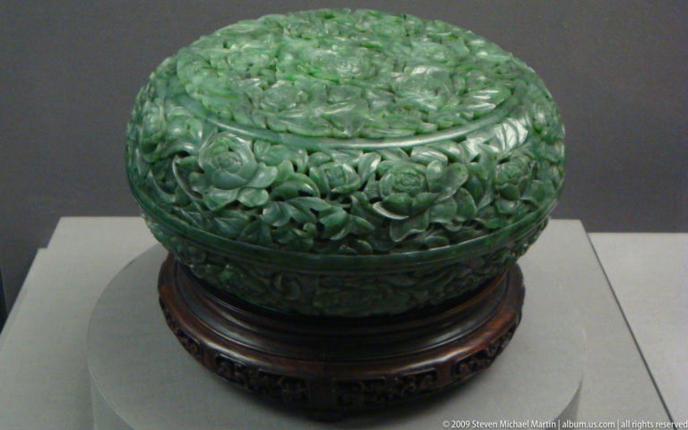 Round deep green jade covered box with dense high-relief floral carving on a wooden stand.