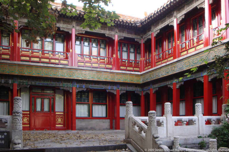 Two-story red residential courtyard with red colonnades, upper gallery, blue-green painted frieze, and white stone balustrade.