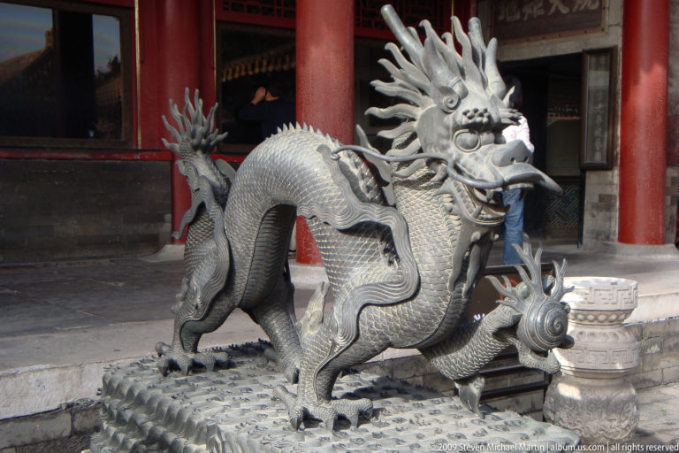 Cast-metal dragon sculpture with raised head, antler-like horns, scales, and cloud-supported forelimb in a Forbidden City courtyard.