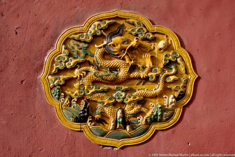 Glazed ceramic relief plaque with a five-clawed yellow dragon among green clouds, framed by a scalloped yellow border on a red wall.