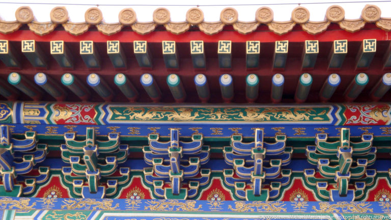 Close-up of painted blue-green dougong brackets under palace eaves, with repeated gold wan symbols on red beam ends.