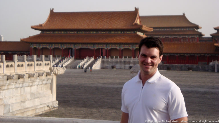 Male visitor in a white shirt standing in the Forbidden City courtyard with a large red-and-gold ceremonial hall on a stone terrace behind him.
