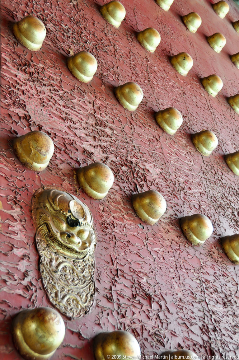 Close-up of gilded door studs and a lion-head boss on a cracked vermilion gate in the Forbidden City.