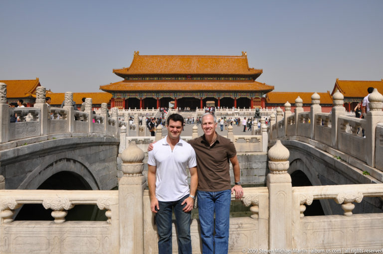 Two male visitors standing on a white stone bridge in front of a large yellow-roofed ceremonial hall in the Forbidden City.