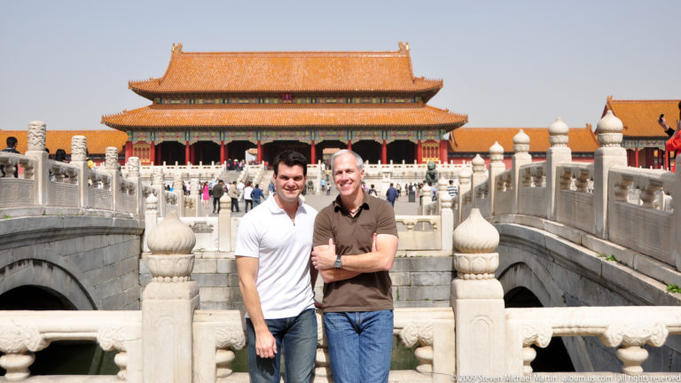 Two male visitors stand on a white stone bridge before a large yellow-roofed ceremonial hall in the Forbidden City.