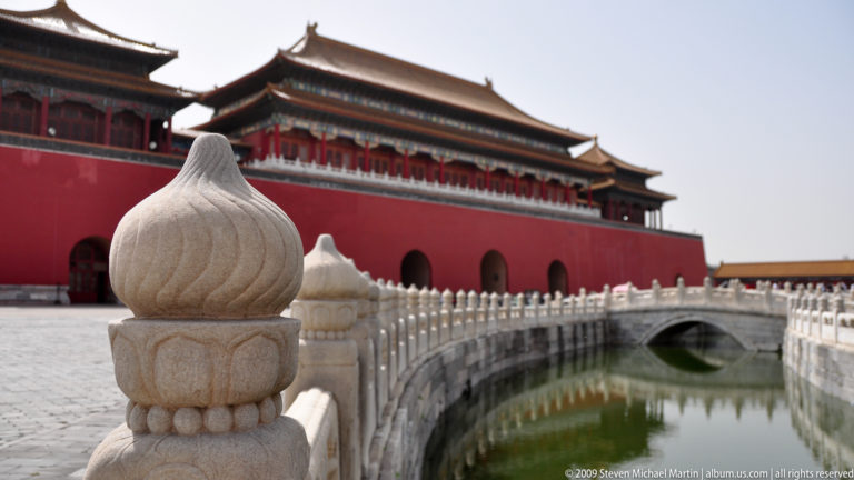 Shallow-focus view across a curving stone bridge toward the red wall and arched opening of the Meridian Gate reflected in the Inner Golden Water River.