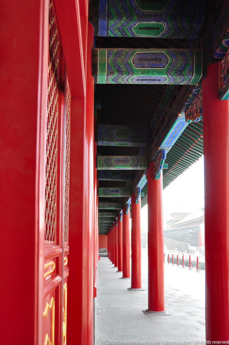 Red-columned covered corridor in the Forbidden City with painted beams and ceiling panels receding into the distance.