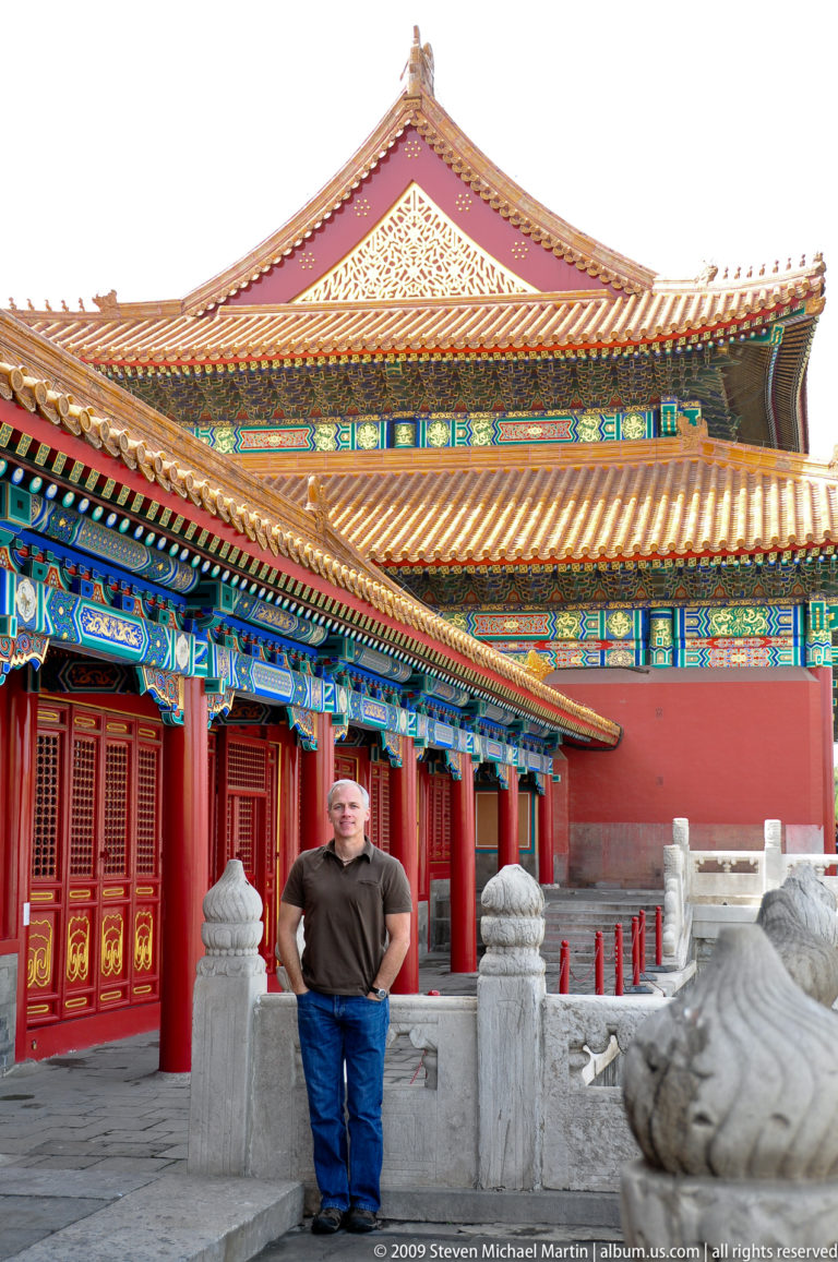 Male visitor standing in a Forbidden City palace courtyard beneath decorated eaves and in front of layered yellow roofs.