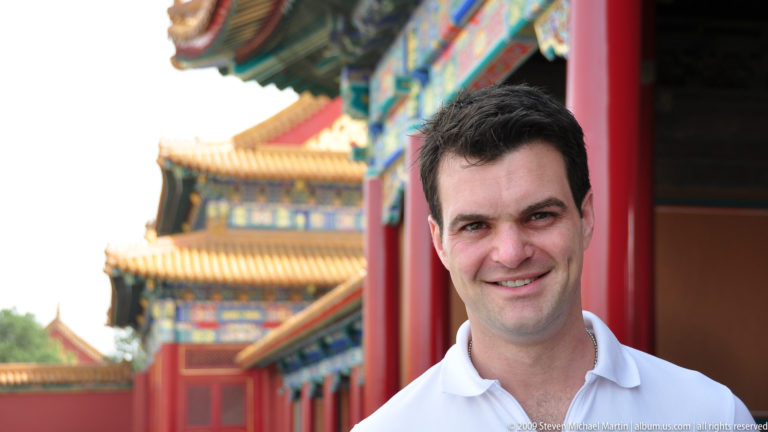 Head-and-shoulders portrait of a dark-haired male visitor in a Forbidden City courtyard, with red columns and yellow-roofed palace buildings softly out of focus behind him.