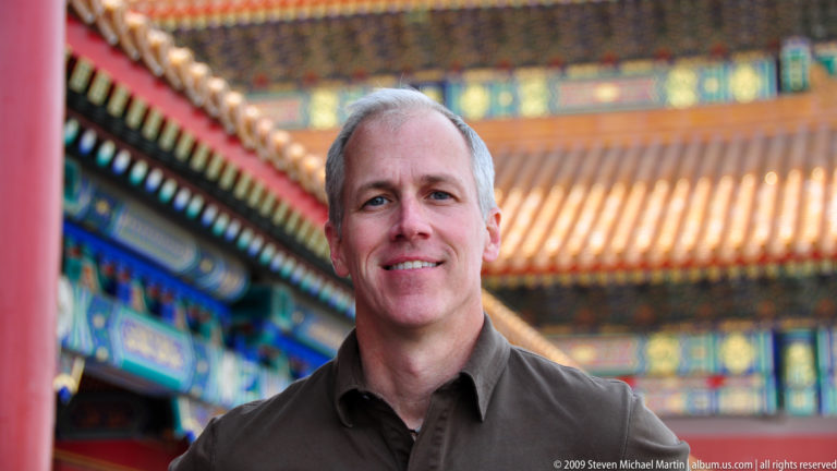 Gray-haired male visitor in sharp focus with blurred red columns, painted brackets, and yellow roofs of the Forbidden City behind him.