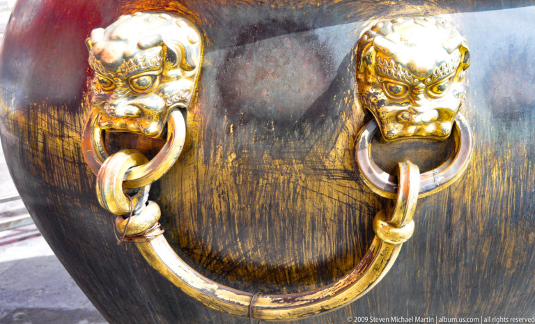 Close-up of two gilded beast-mask ring handles on a dark curved bronze vessel wall, each mask gripping a thick circular ring.