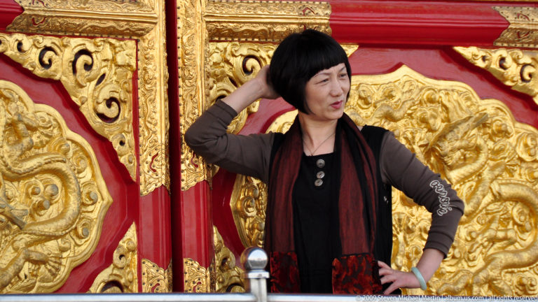 Woman posing with one hand raised in front of richly carved red and gold gilded doors at the Forbidden City.