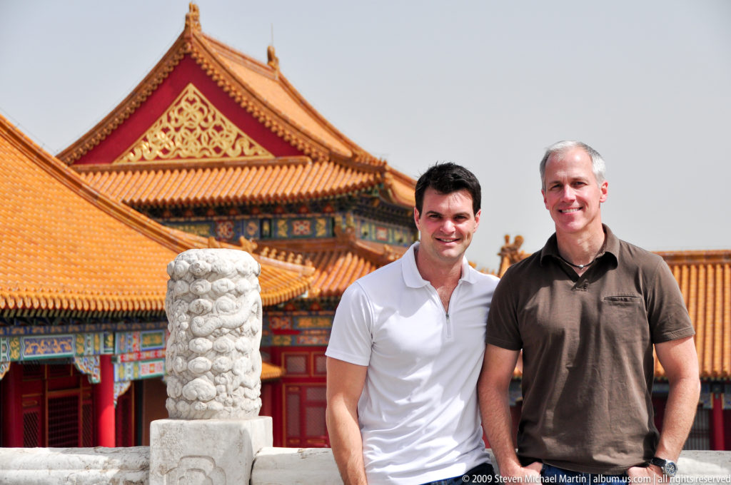 Two male visitors stand together smiling in front of yellow roofs, red walls, and carved stone details at the Forbidden City.
