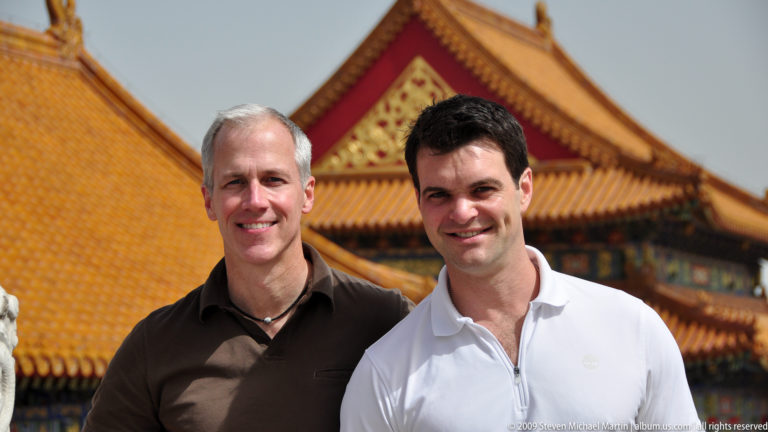 Two male visitors smiling and standing together with the yellow roofs and red walls of the Forbidden City behind them.
