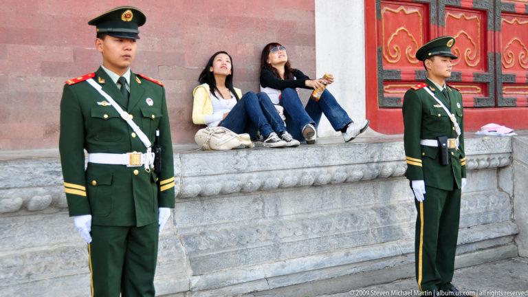 Two modern Chinese honor guards in green uniforms and peaked caps stand at attention while two tourists sit on a stone ledge between them in the Forbidden City, Beijing.