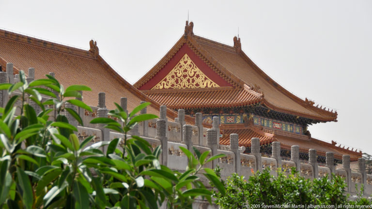 Oblique view of a yellow-roofed palace hall with gold gable ornament, partly screened by green foliage and a white stone terrace.