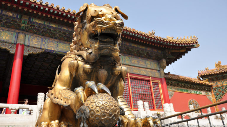 Low-angle close-up of a gilt-bronze male guardian lion with an open mouth and one paw resting on a patterned brocade ball.