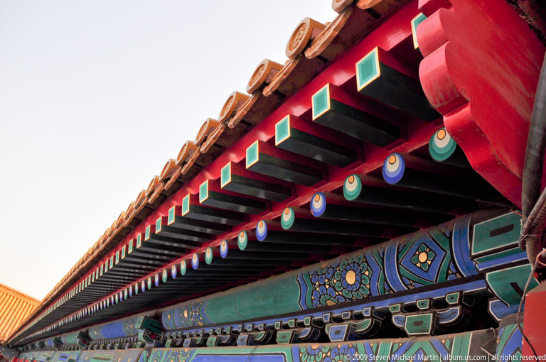 Diagonal view of the underside of a Forbidden City palace roof, showing repeated red rafters and a blue-green floral painted beam.