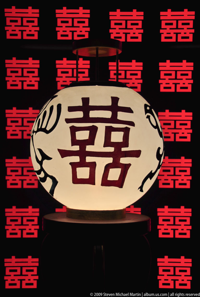 Cream lantern with large dark red 囍 double-happiness character against a patterned red-and-black background of smaller 囍 symbols.