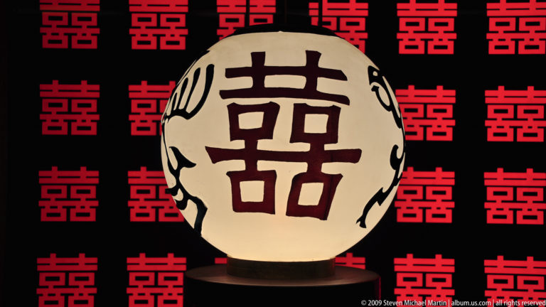 Centered cream lantern with the 囍 double-happiness character, set before a backdrop of repeated red symbols.
