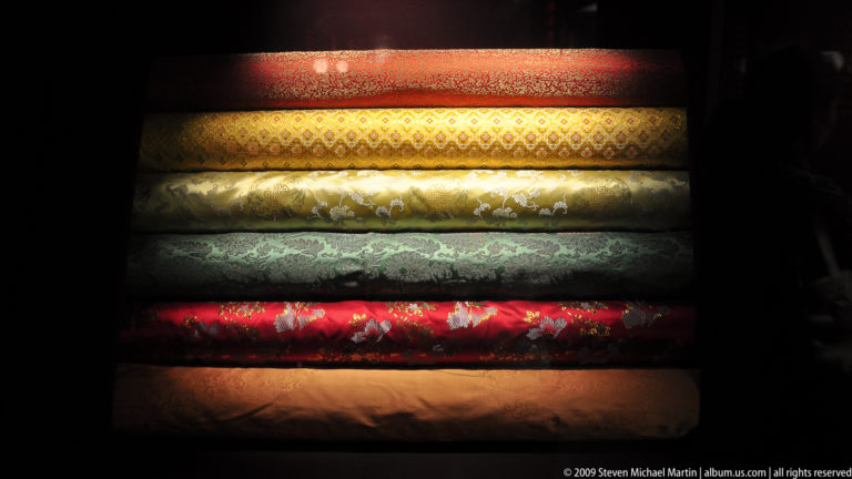 Horizontal museum display of stacked rolls of red, gold, green, and blue-green silk brocade, each with a distinct woven floral or geometric pattern.