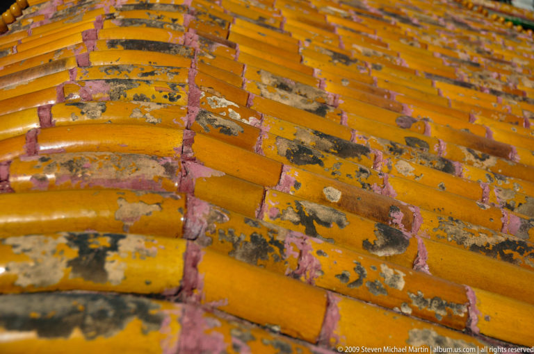 Close-up of weathered yellow-glazed roof tiles showing flaking glaze and exposed reddish material beneath.