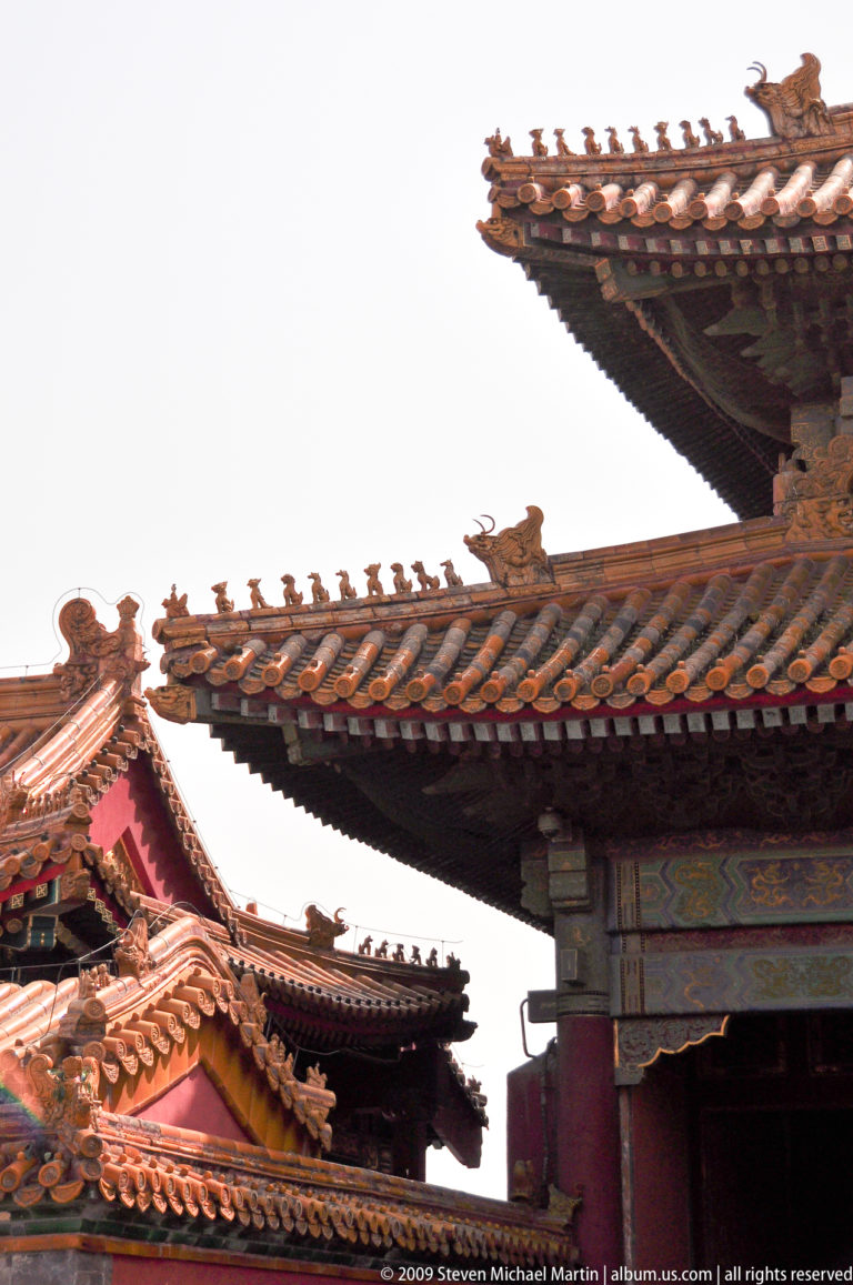 Upward view of overlapping palace eaves with small roof figurines along one ridge against a bright sky.