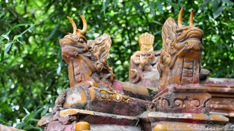 Close-up of three weathered glazed-ceramic dragon or chiwen ridge ornaments on a garden roof, with worn glaze exposing reddish ceramic beneath.