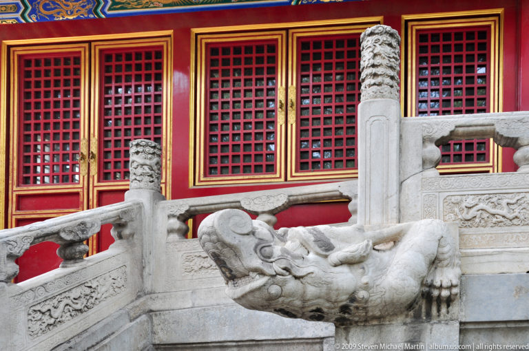 Close view of a carved stone dragon-head waterspout projecting from a white palace terrace with red lattice windows behind.