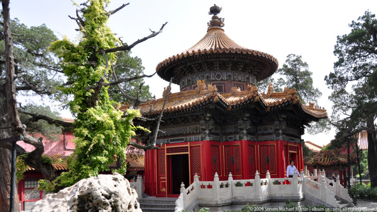 Round red pavilion with a conical glazed roof surrounded by rocks and trees in the Imperial Garden of the Forbidden City.