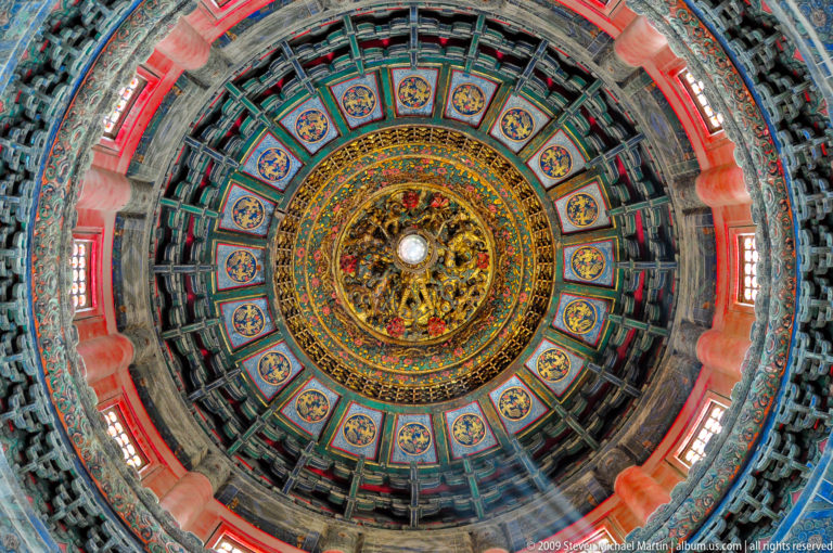 Upward view of a radial caisson ceiling with blue-green coffers and a central gold dragon medallion in an Imperial Garden pavilion.