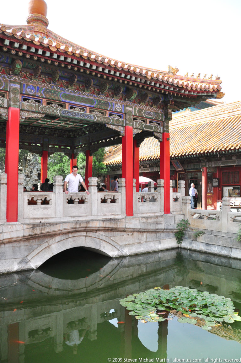 Visitor standing in a red-columned pavilion over a pond beside a white stone arch bridge in the Imperial Garden of the Forbidden City.