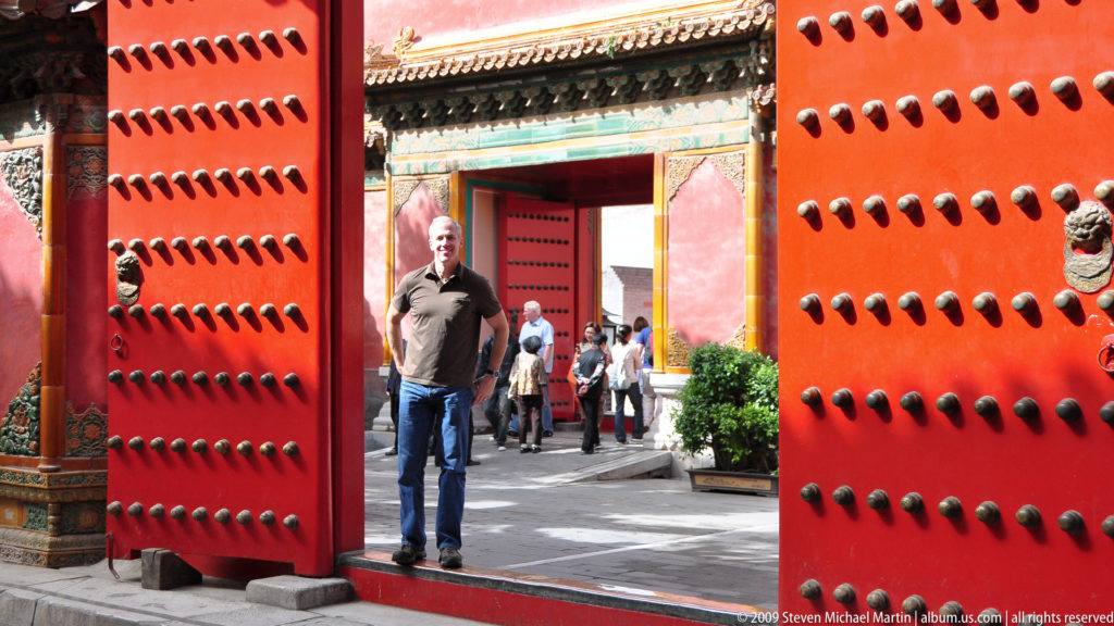 Full-length portrait of a male visitor standing inside an open red studded palace gate, with a second decorated doorway visible in line behind him.