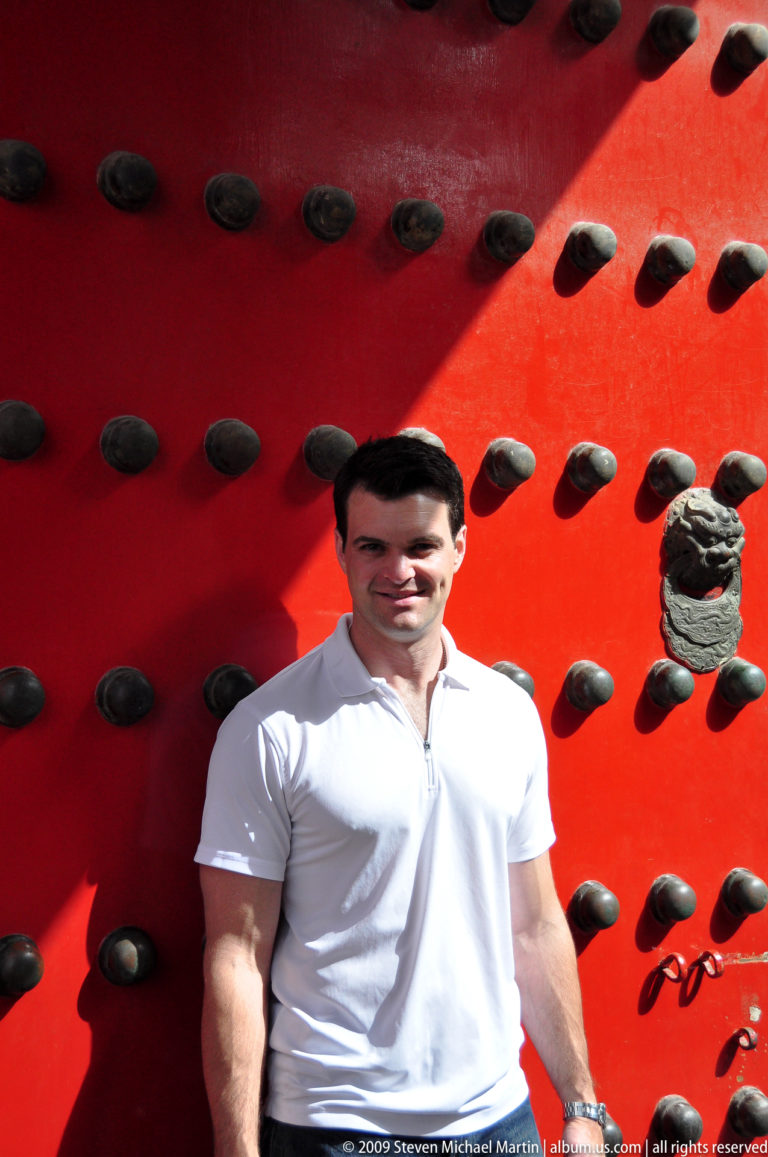 Male visitor in front of a bright red studded door with a sculpted lion-head fitting beside him.