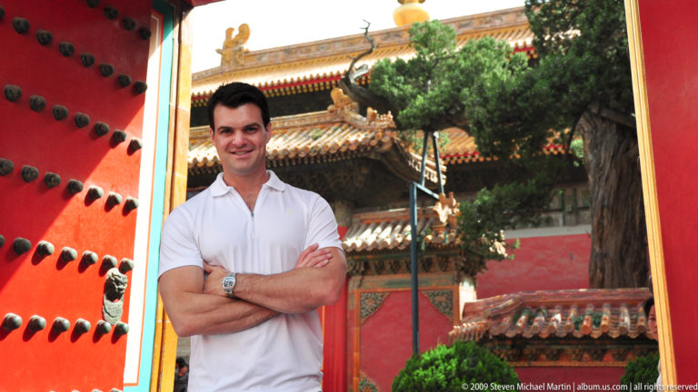 Male visitor with arms crossed standing beside a red palace gate and an evergreen tree in the Forbidden City.